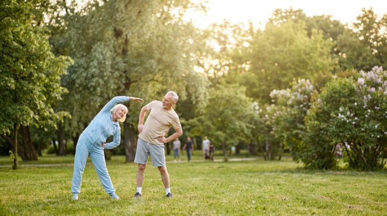 Un couple de personnes âgées faisant des étirements ensemble sur une pelouse verdoyante dans un parc ensoleillé.
