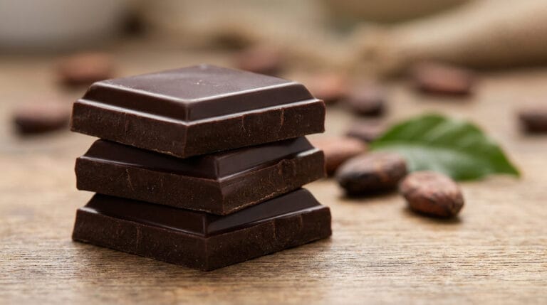 Trois carrés de chocolat noir empilés sur une table en bois avec des fèves de cacao en arrière-plan.
