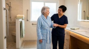 Une aide-soignante accompagne une femme âgée en peignoir dans une salle de bain accessible et sécurisée.