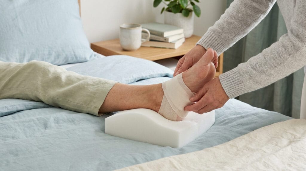Une personne ajuste un bandage sur le talon d'un patient reposant sur un coussin en mousse dans un lit.