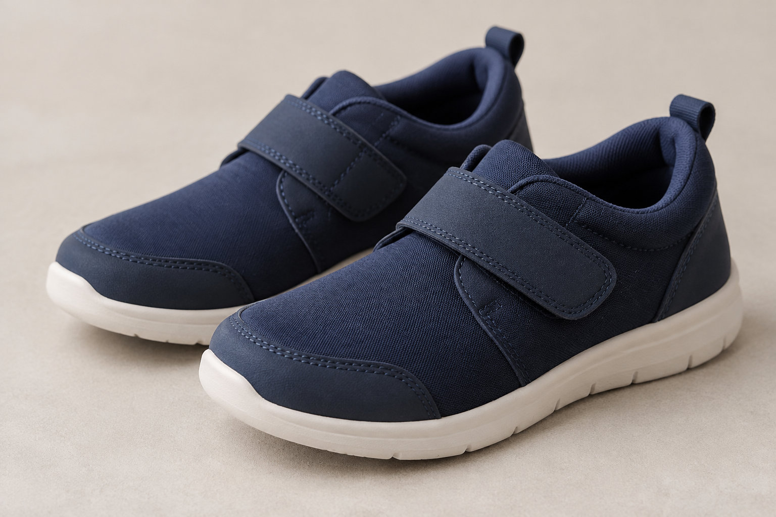 Deux chaussures de sport bleu marine à fermeture autoagrippante et semelles blanches sont posées sur une surface beige.